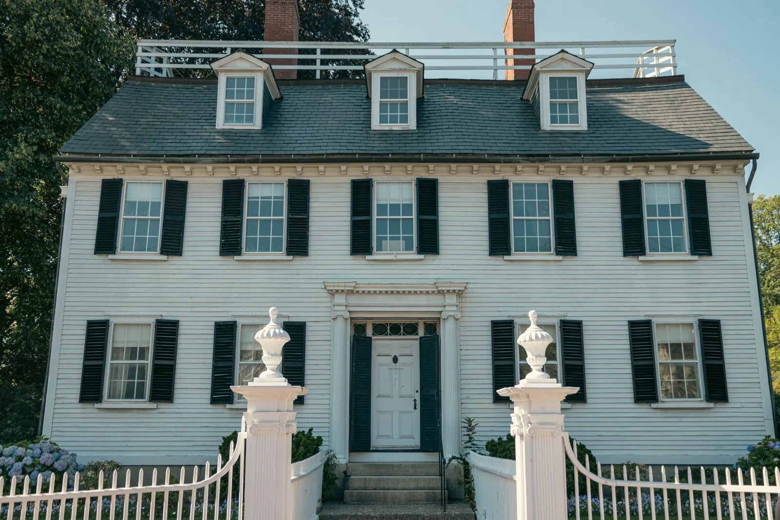 White clapboard colonial with black shutters, brick chimneys and a picket fence