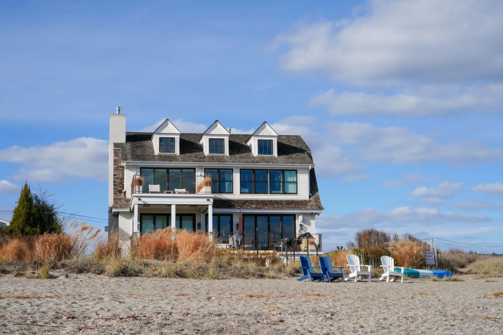 Gray shingle beach house with dormers and a chimney above sandy dunes